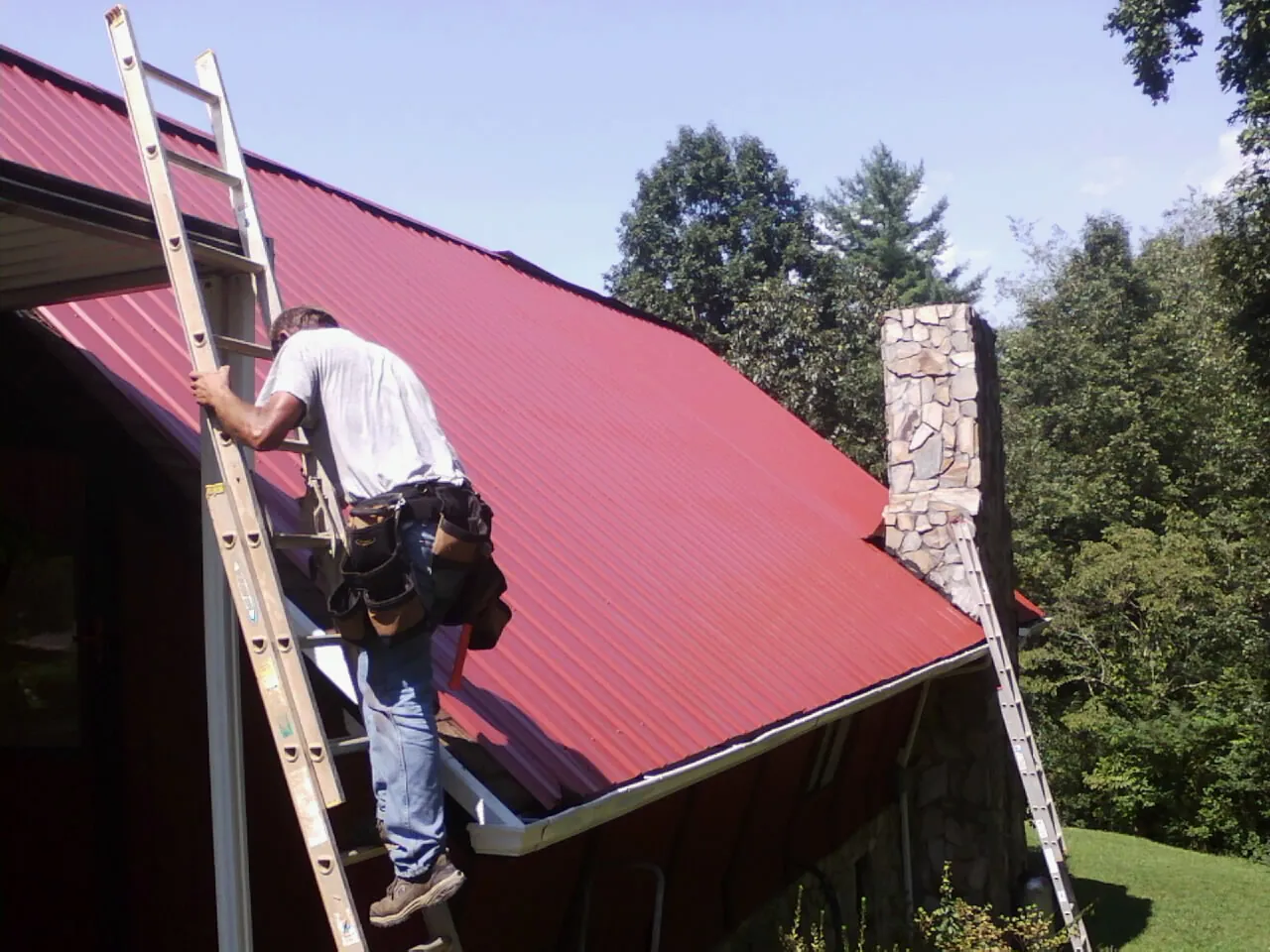 Roofer on a ladder beside a red metal roof and stone chimney — Soffit & Fascia Repair by Roofing Brothers in Sauk Centre